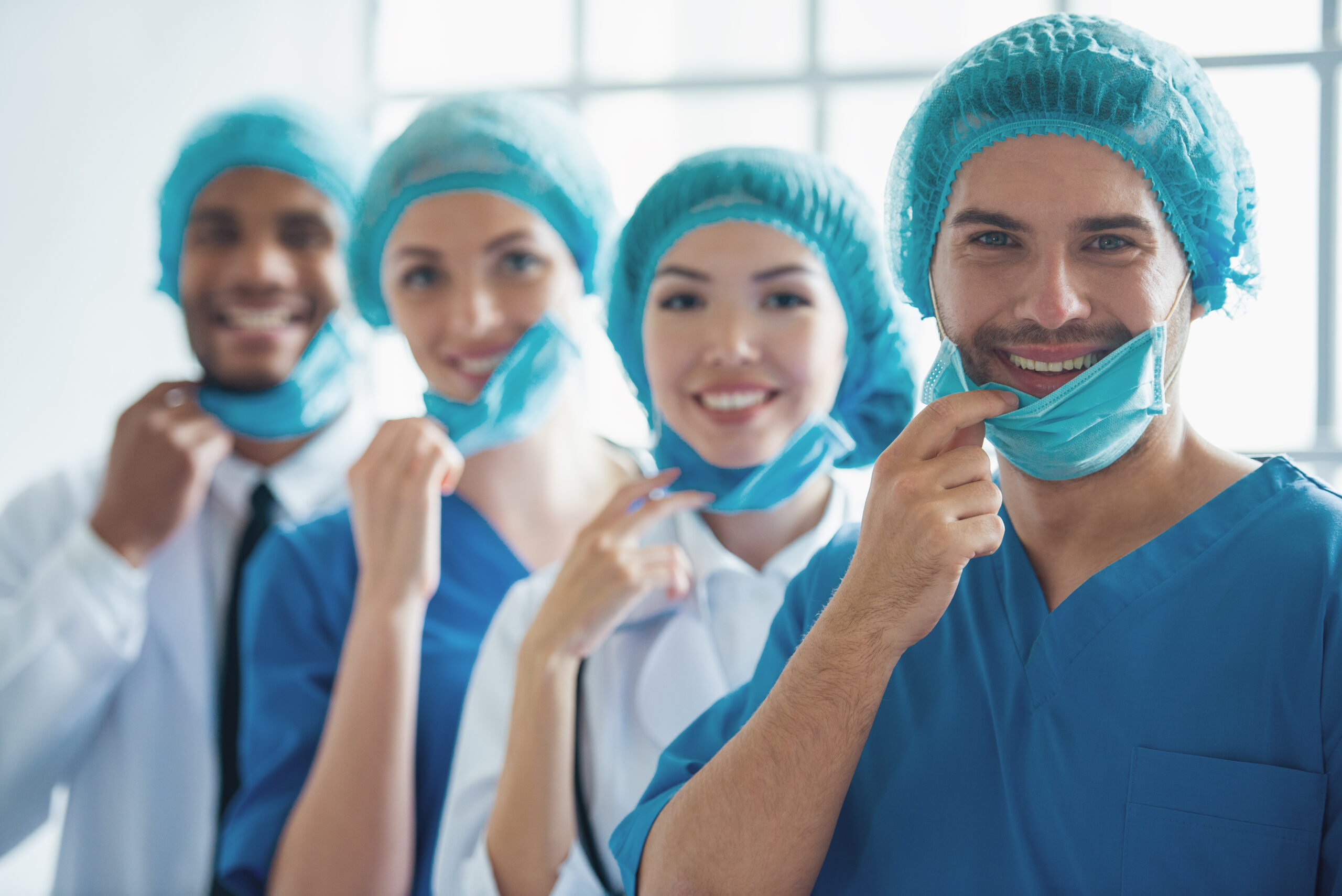 Medical doctors of different nationalities and genders in caps are pulling masks, looking at camera and smiling