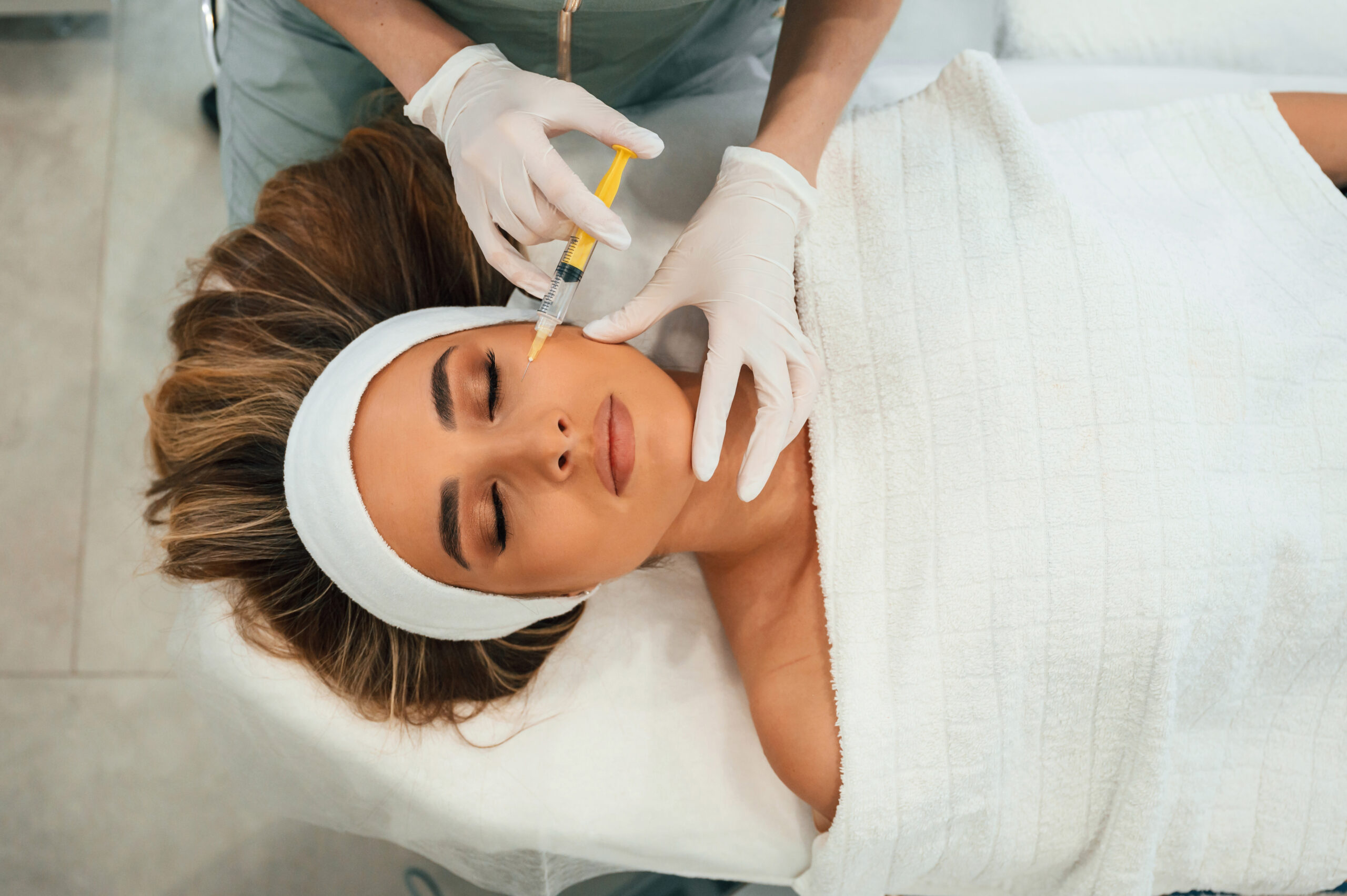 Yellow colored syringe, making injection. Woman face getting facial care by beautician hands at spa salon.